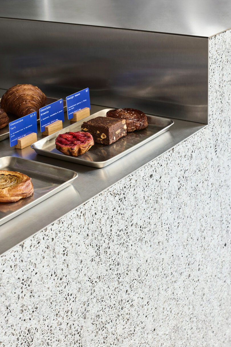 A display counter with metal trays holding assorted pastries and cakes, each labeled with blue cards, set on a terrazzo surface.