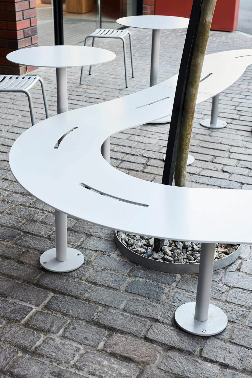 Curved white outdoor table with slot openings built around a tree on a cobblestone surface, accompanied by metal stools.