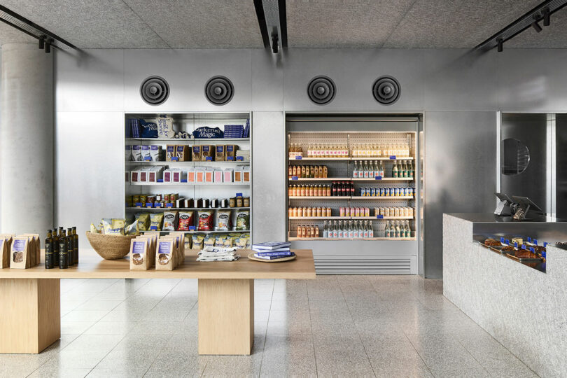 Minimalist shop interior with shelves of packaged foods, a refrigerated display stocked with drinks, and a wooden table displaying various products in the foreground.