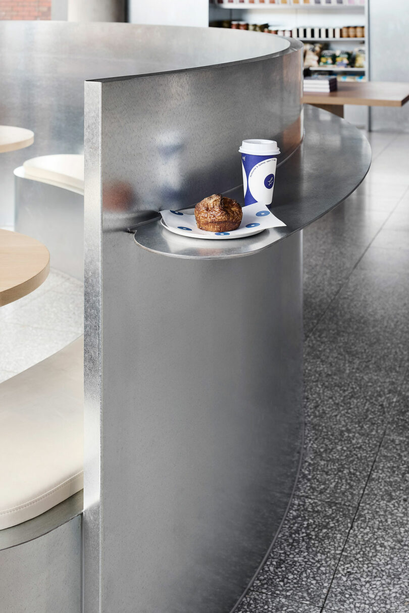 A paper coffee cup and a pastry on a napkin sit on a small ledge of a curved metallic partition in a modern cafe interior.