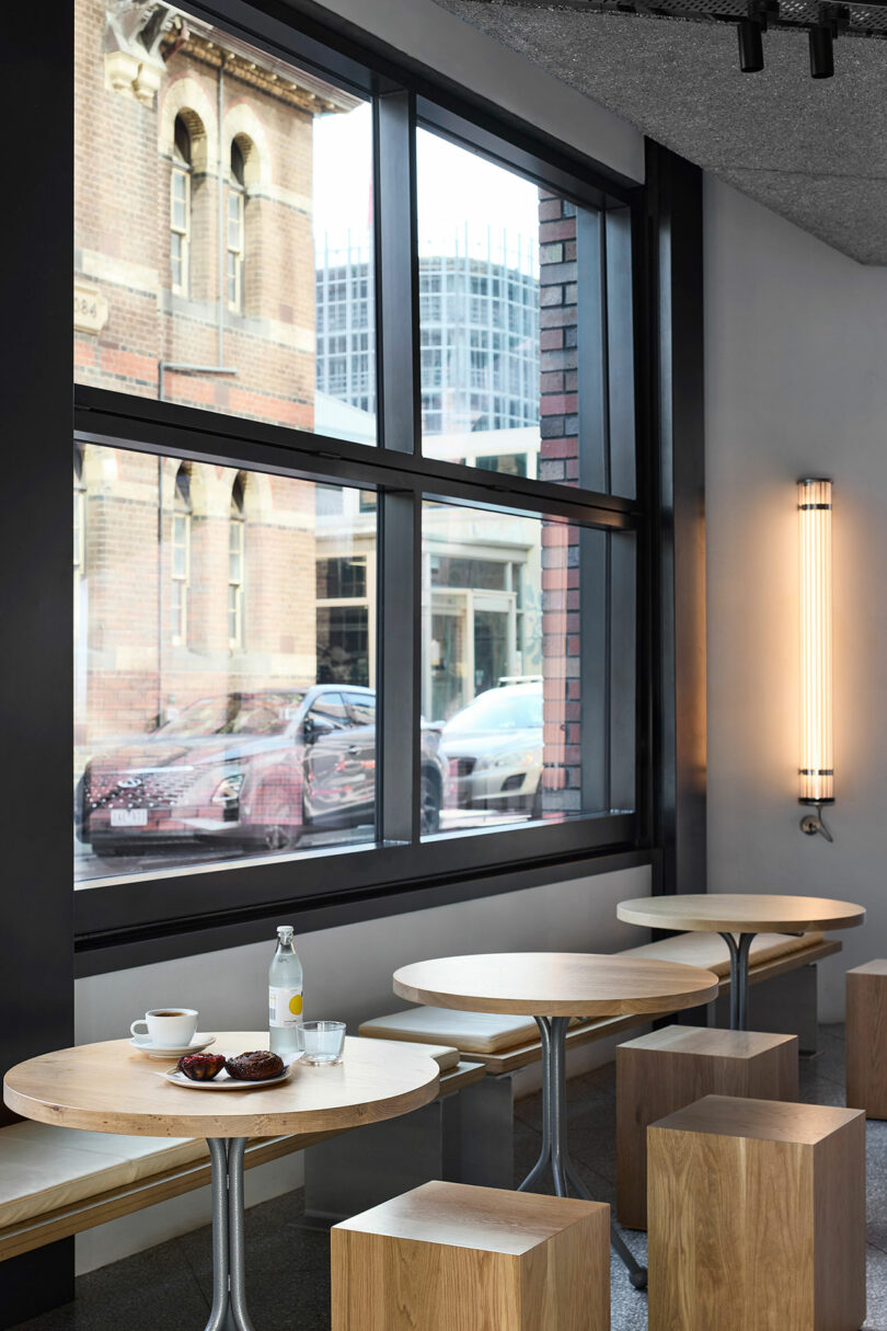 Minimalist cafe interior with round wooden tables, wooden stools, a cup of coffee, plate with pastry, glass of water, and a bottle, beside large windows showing parked cars outside.