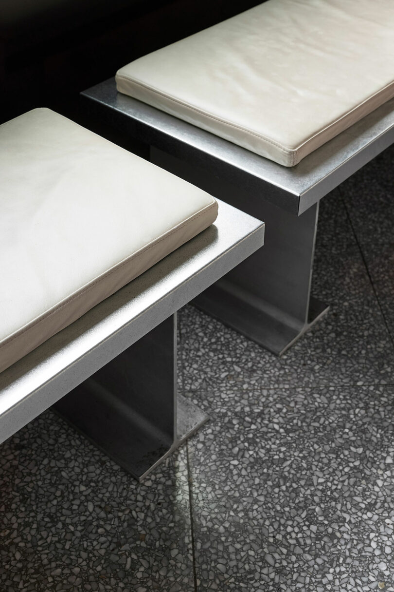 Two metal benches with white seat cushions are positioned on a terrazzo floor.