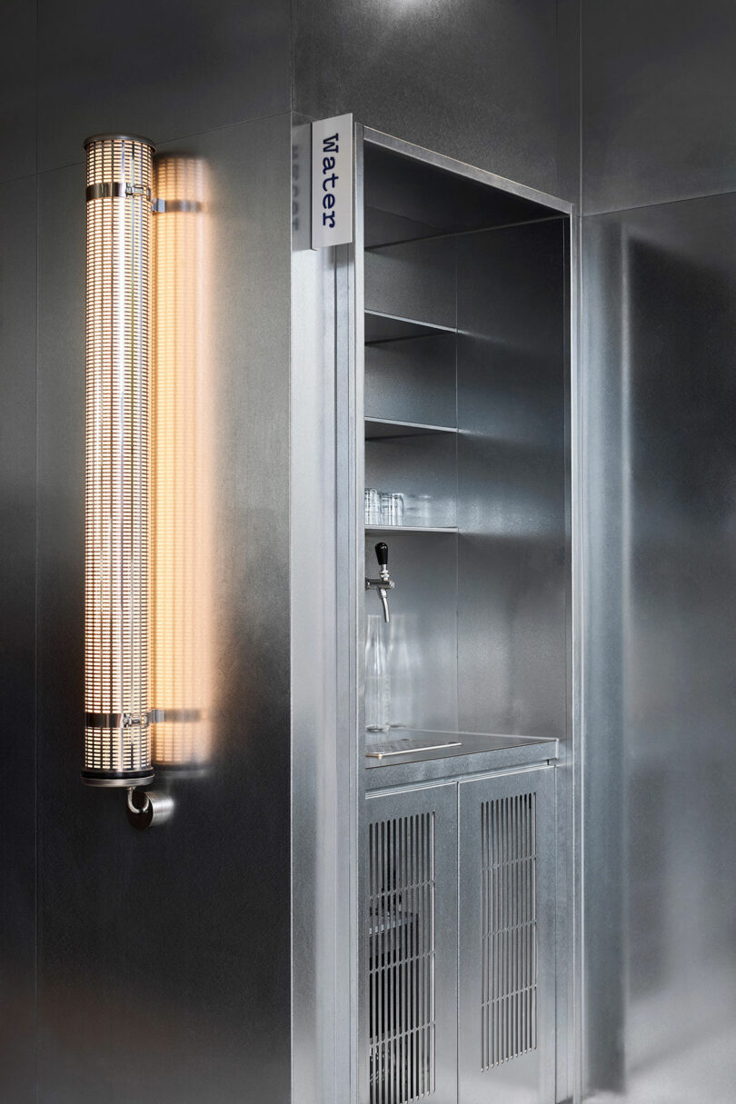 A metallic water station with a glass bottle and a faucet is set into a wall, next to a vertical, cylindrical light fixture. A sign labeled