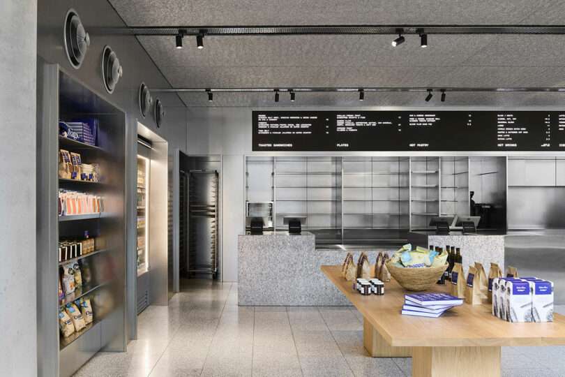 Modern bakery interior with metal shelves, a wooden display table with packaged goods, a refrigerated section, and digital menu boards above a granite counter.