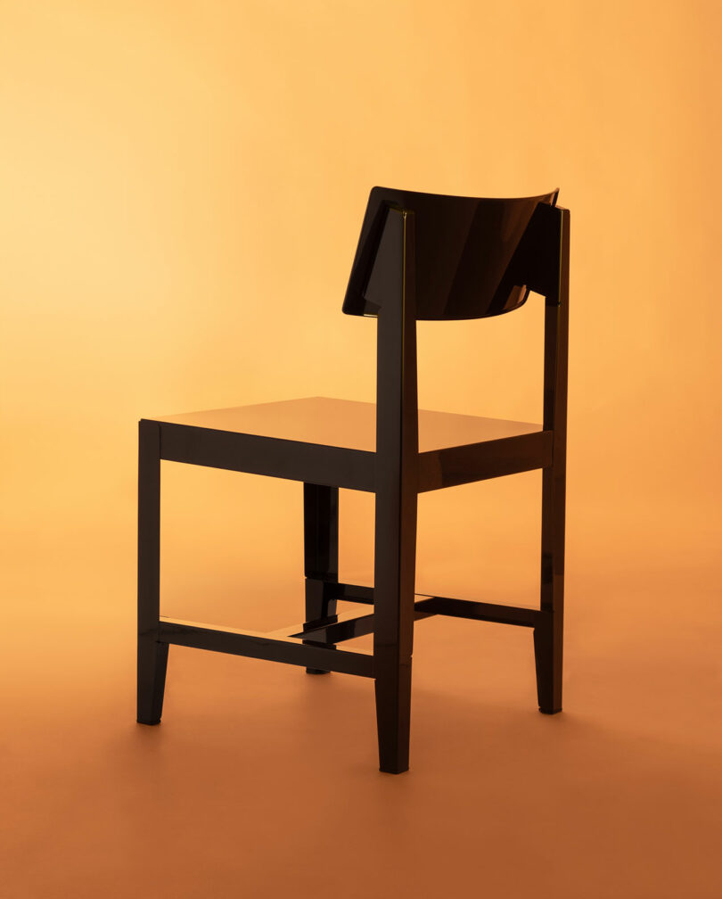 A black, modern chair with a solid backrest and four straight legs, placed on an orange background.