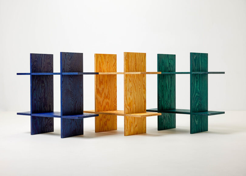 Three freestanding shelving units in blue, yellow, and green wood, each with intersecting vertical and horizontal panels, set against a plain white background.