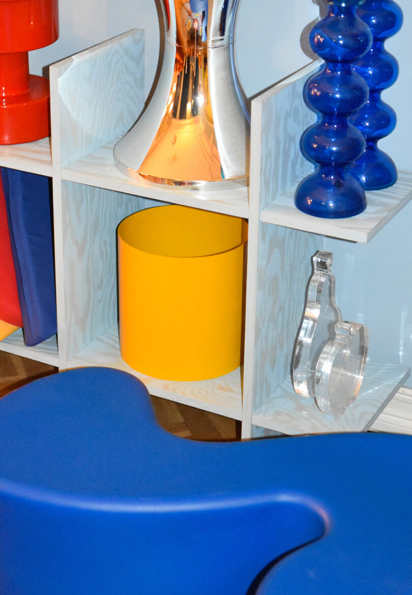 A light blue wooden shelf displays a yellow cylinder, blue and red sculptural objects, a mirrored surface, and a clear abstract form. A blue curved object is in the foreground.