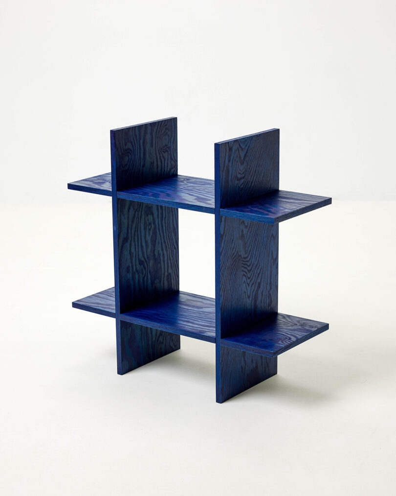 A minimalist, modular blue wooden shelf with two horizontal and two vertical panels, forming four open compartments, set against a plain white background.