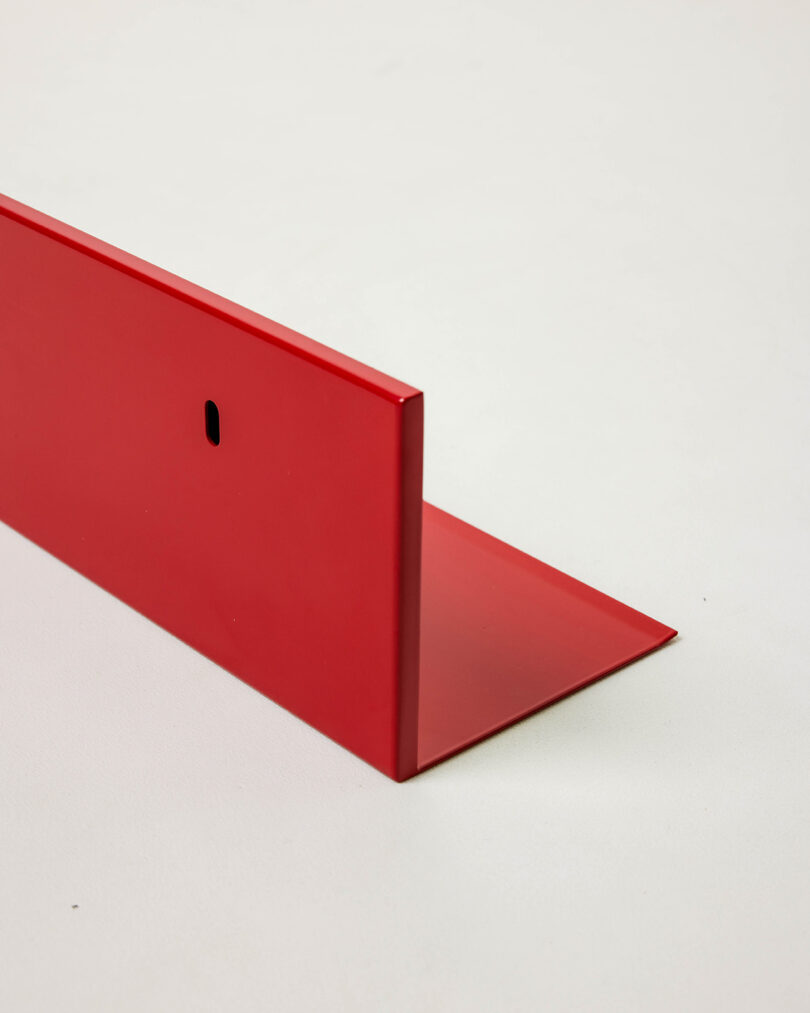 A red L-shaped metal bracket with a small oval hole near one edge, placed on a plain white surface.
