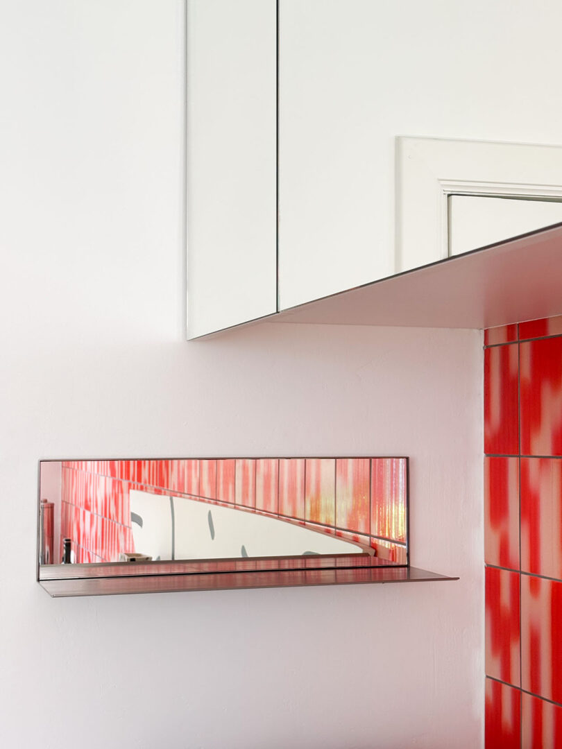 A rectangular wall-mounted mirror shelf reflects a red and white geometric-patterned wall in a corner of a modern room.