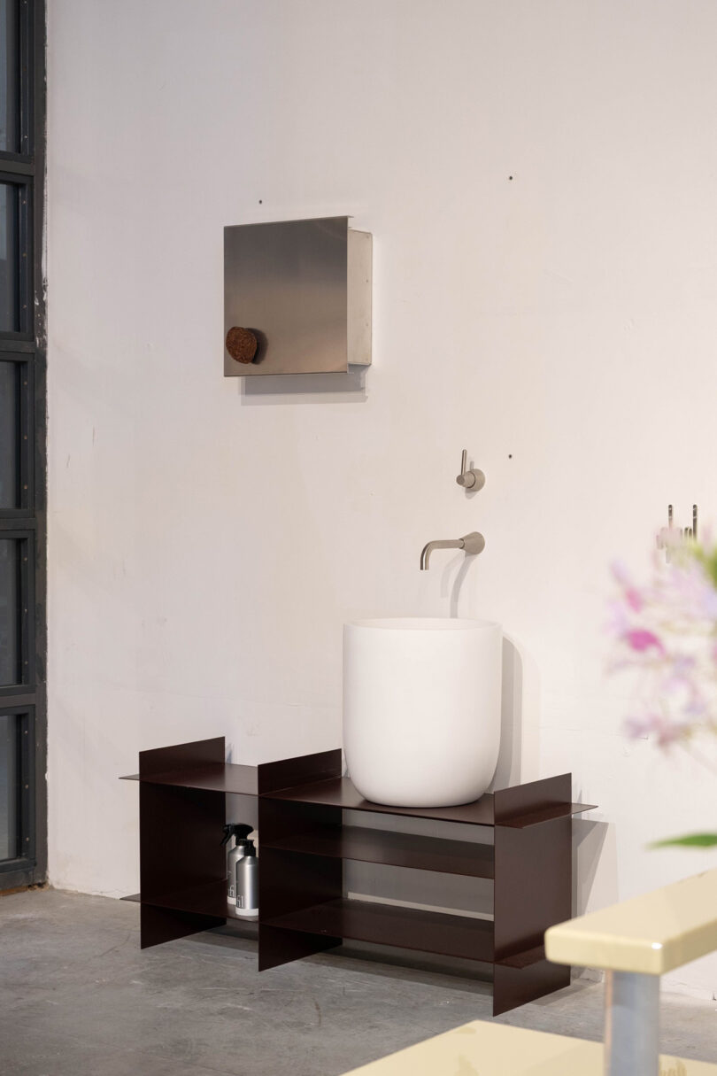 A modern bathroom sink with a round white basin sits on a dark open shelf unit; a wall-mounted faucet and a metal cabinet are installed above.