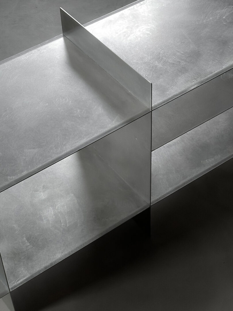 Two minimalist metallic shelves intersect at right angles, forming a simple geometric structure on a smooth, light-colored surface.