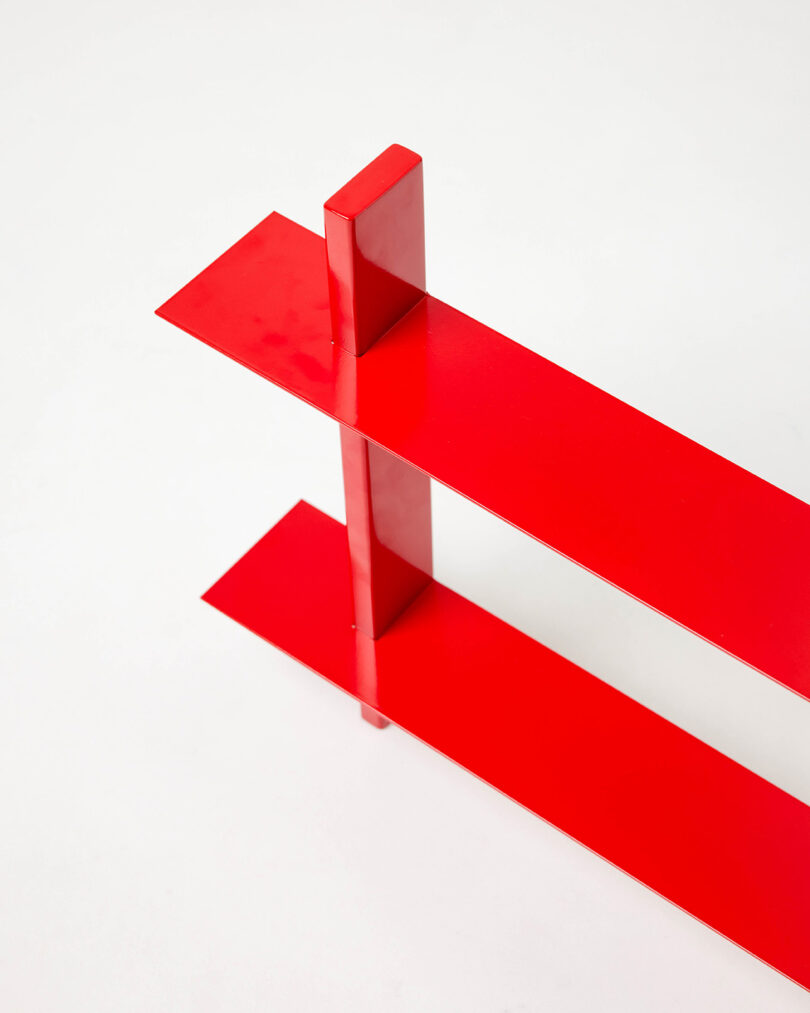 Two glossy red rectangular metal shelves intersect perpendicularly, forming a minimalist geometric structure against a plain white background.