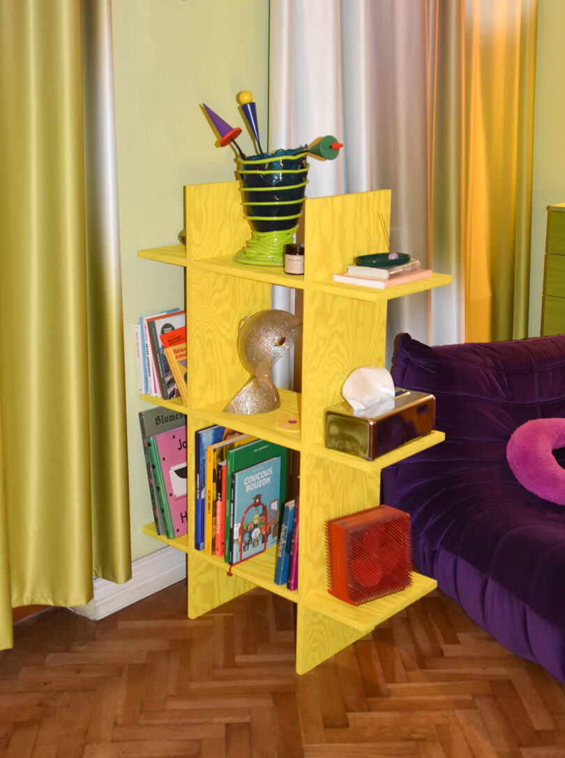 A yellow wooden shelf holds colorful books, a green vase with utensils, a gold mannequin head, a tissue box, and other decorative items next to a purple sofa and green curtains.