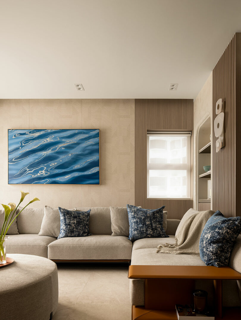 A modern living room with a beige sectional sofa, blue patterned pillows, wall art depicting water, a round ottoman, a side table, and a window with a roller shade.
