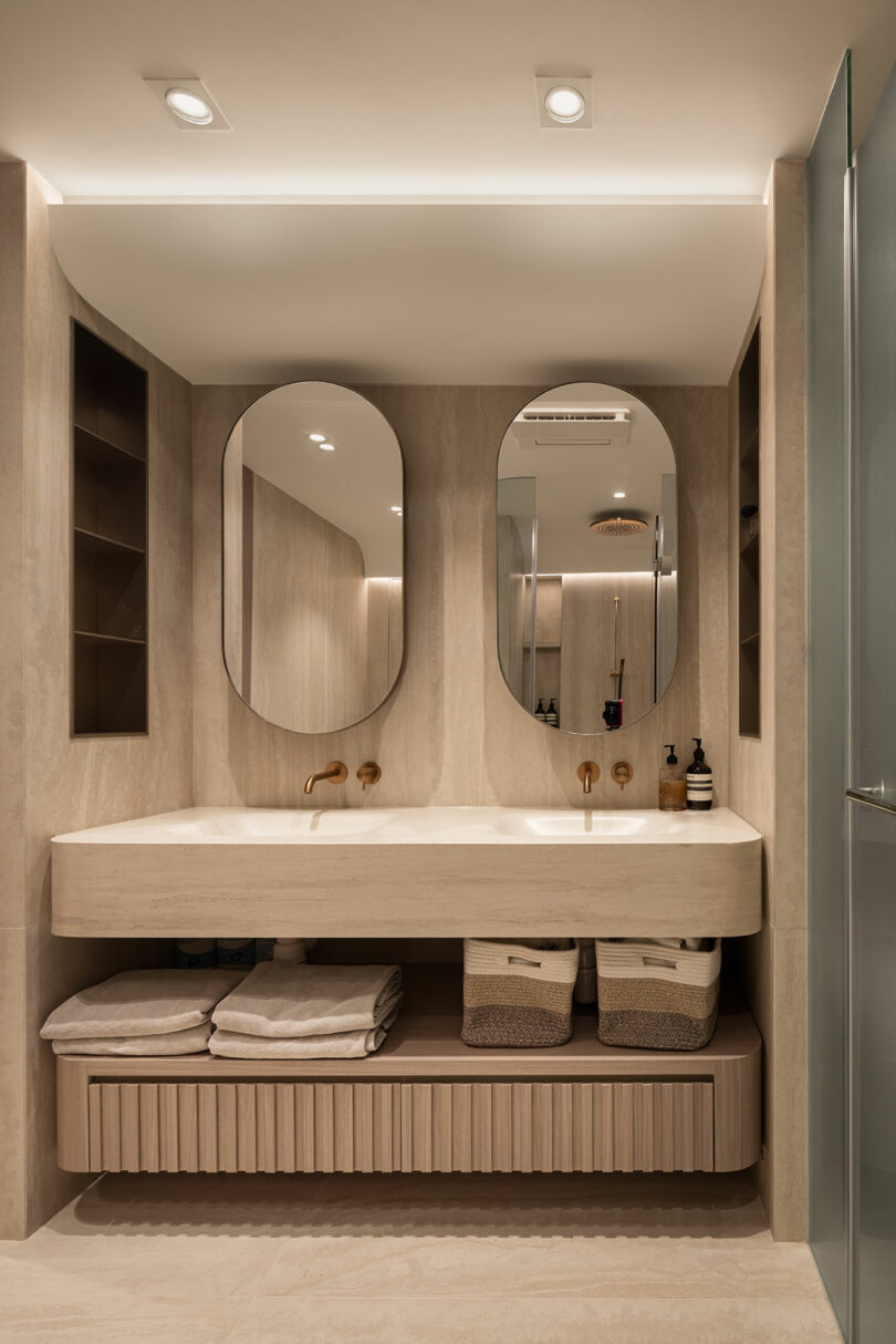 Modern bathroom with double oval mirrors, a beige stone sink, open shelving, two baskets underneath, and neatly folded towels on a wooden shelf.