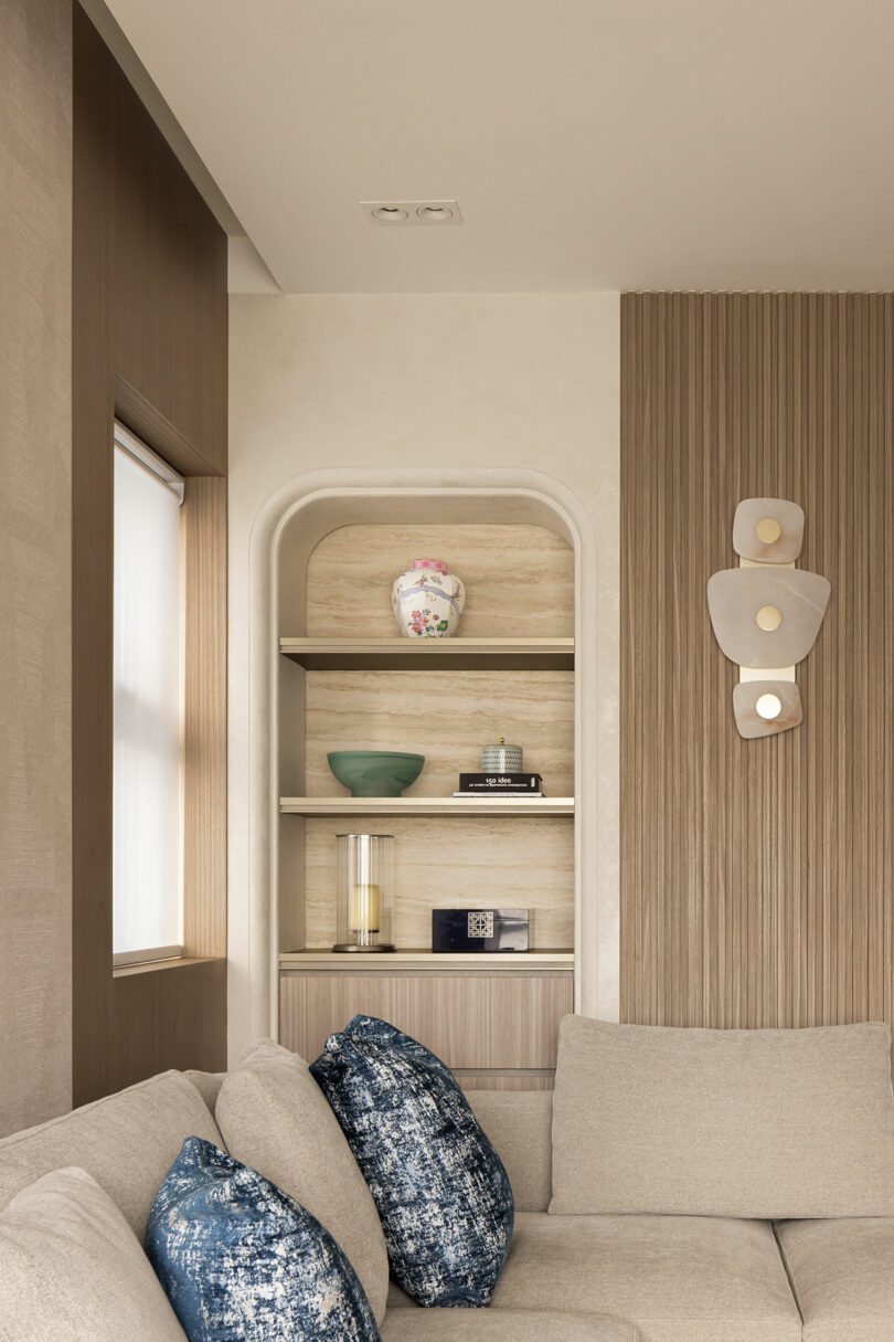 A modern living room corner with a beige sofa, blue-patterned pillows, built-in shelving with decorative items, and a wall-mounted sculptural light fixture.