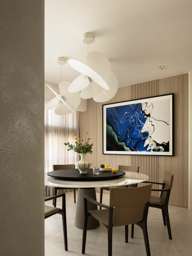Modern dining area with a round table, four chairs, pendant lights, a vase of yellow flowers, and abstract art on a wood-paneled wall.
