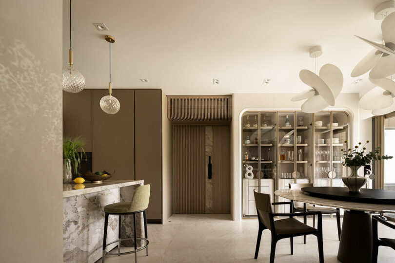 Modern kitchen and dining area with marble surfaces, glass-fronted cabinet, olive green accents, and sculptural white ceiling lights.