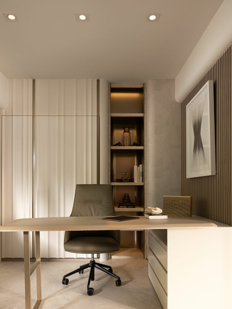 Minimalist office with a beige desk, swivel chair, built-in shelves with books and decor, wall art, and soft ambient lighting.