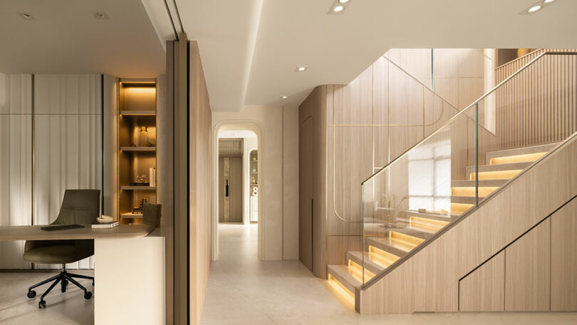 Modern interior with a built-in desk, office chair, shelves, and a wooden staircase with illuminated steps and glass railing, featuring neutral tones and minimalist design.
