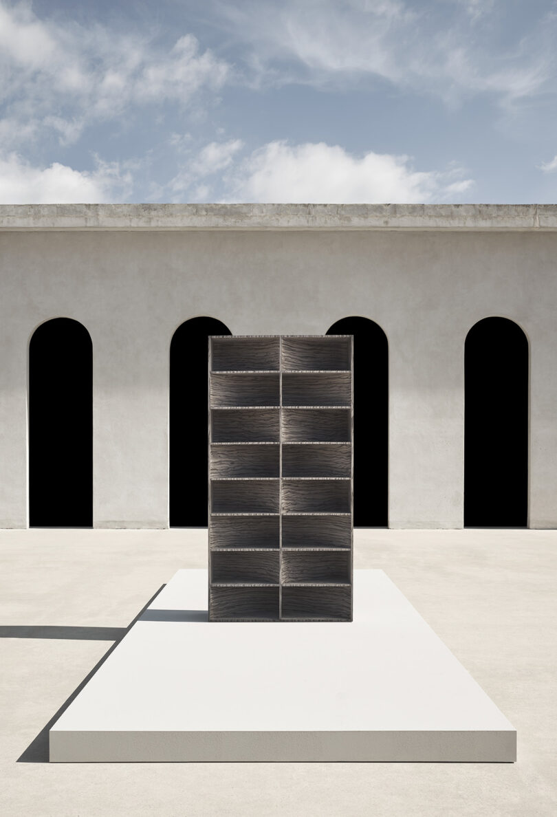 A rectangular shelving unit with twelve open compartments stands on a white platform in front of a building with three tall arched doorways.