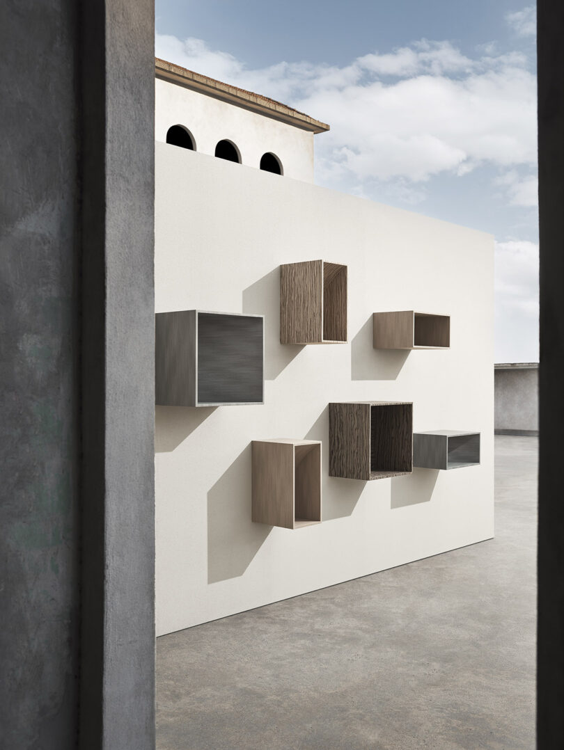 A white wall outdoors with various rectangular wooden and metal shelves mounted in an irregular pattern, viewed between concrete structures under a partly cloudy sky.