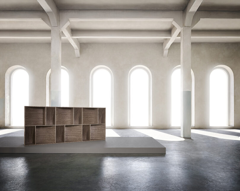 A modern, wooden cabinet sits on a platform in a spacious room with high ceilings and tall, arched windows letting in natural light.