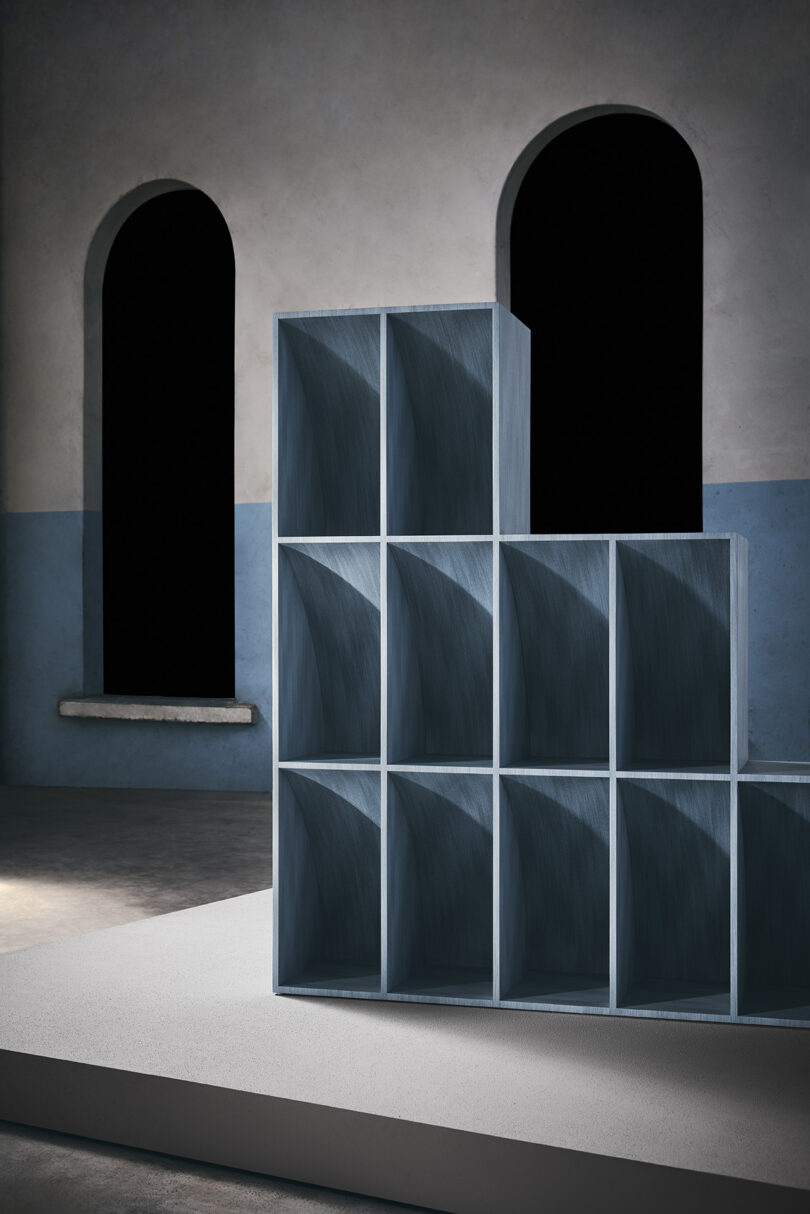 A staggered, blue-gray cubby shelf stands on a platform in front of two arched windows with blacked-out panes in a minimalist, two-tone room.