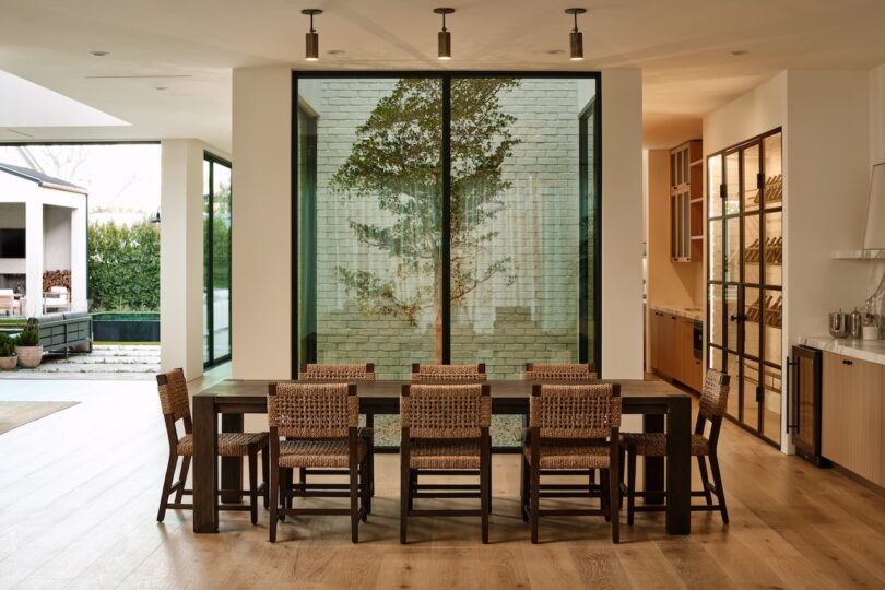 A modern dining area with a wooden table and eight woven chairs, featuring a large window with a tree view and an open layout leading to an outdoor space.