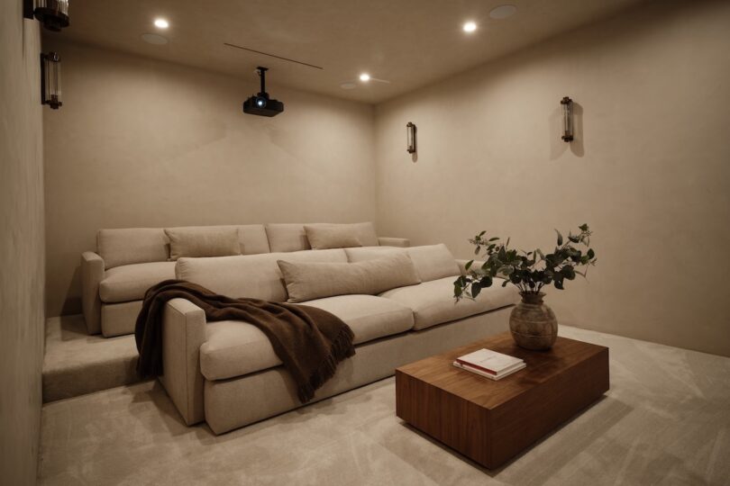 A home theater room with beige walls, tiered cream sofas, a brown throw, a wooden coffee table with books and a vase, and a ceiling-mounted projector.