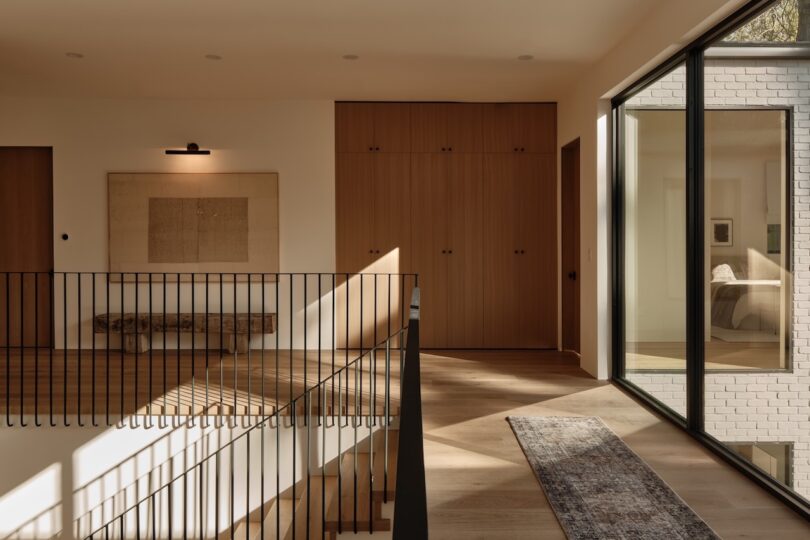 A modern hallway with wooden floors, a metal railing, built-in wooden cabinets, wall art, a bench, and large windows showing a view into a bedroom.