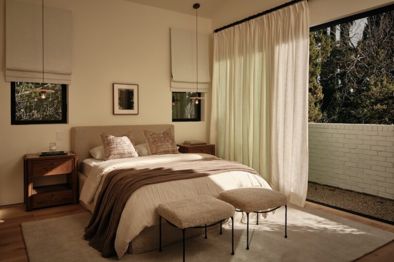 A modern bedroom with a bed, two bedside tables, two benches, large window with sheer curtains, and neutral-toned decor.