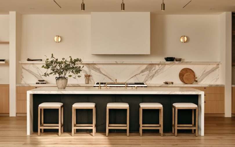 Modern kitchen with a marble island, four stools, minimalist decor, light wood flooring, and built-in shelves. Neutral tones and clean lines create a sleek, contemporary look.