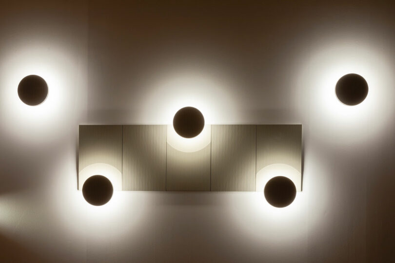 Five circular wall lights evenly spaced above and below a rectangular textured panel, casting a soft glow onto a neutral wall.