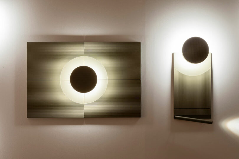 Two modern wall lamps with circular black centers and geometric, ribbed designs emit soft white light against a plain wall.