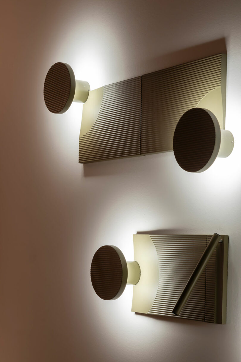 Two modern wall-mounted light fixtures with circular bulbs and textured rectangular bases are illuminated, casting light against a plain white wall.