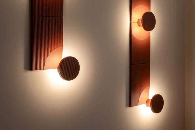 Two modern wall sconces with geometric shapes emit warm light, featuring circular and rectangular copper-colored elements mounted on a light-colored wall.