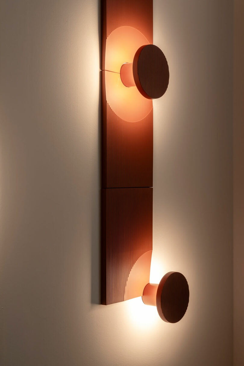 Wall-mounted modern light fixture with two round, ribbed red lamps emitting soft light, attached to a vertical, textured red panel on a neutral wall.