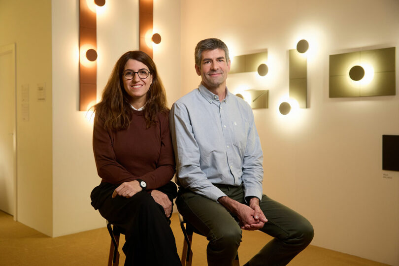 A woman and a man sit side by side on stools in a well-lit room with modern wall lights displayed behind them.