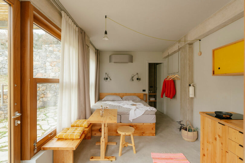 A minimalist bedroom by Meraki Studios featuring a double bed, wooden furniture, large window, small dining area, hanging clothes, and neutral decor.
