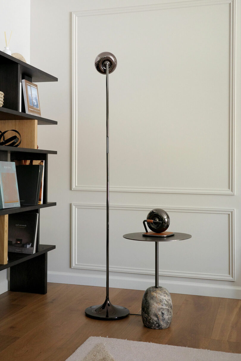Modern living room corner with a tall metallic floor lamp, a small round side table with a decorative object, and a bookshelf with books against a white paneled wall and wooden floor.