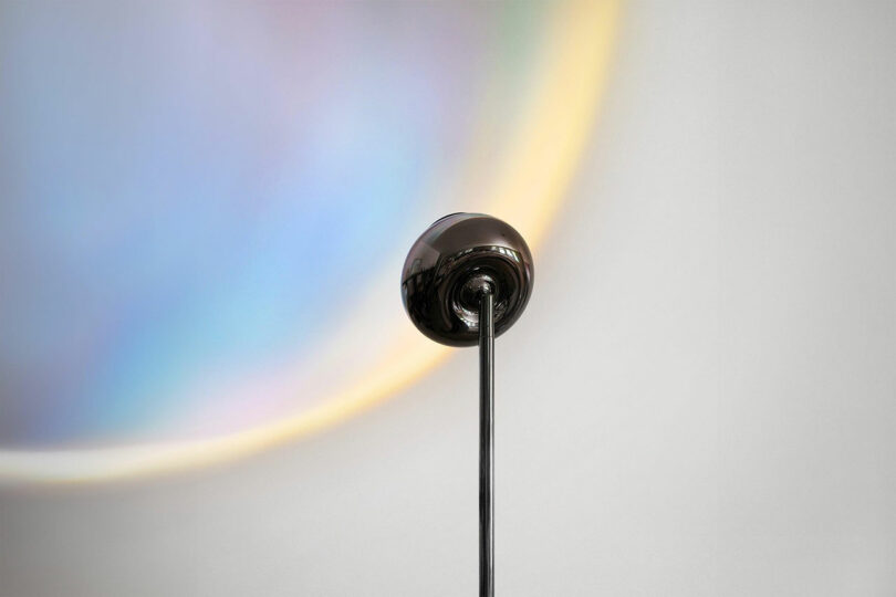 A metallic lamp head on a slim pole stands against a plain wall, casting a soft, circular rainbow-like light pattern in the background.
