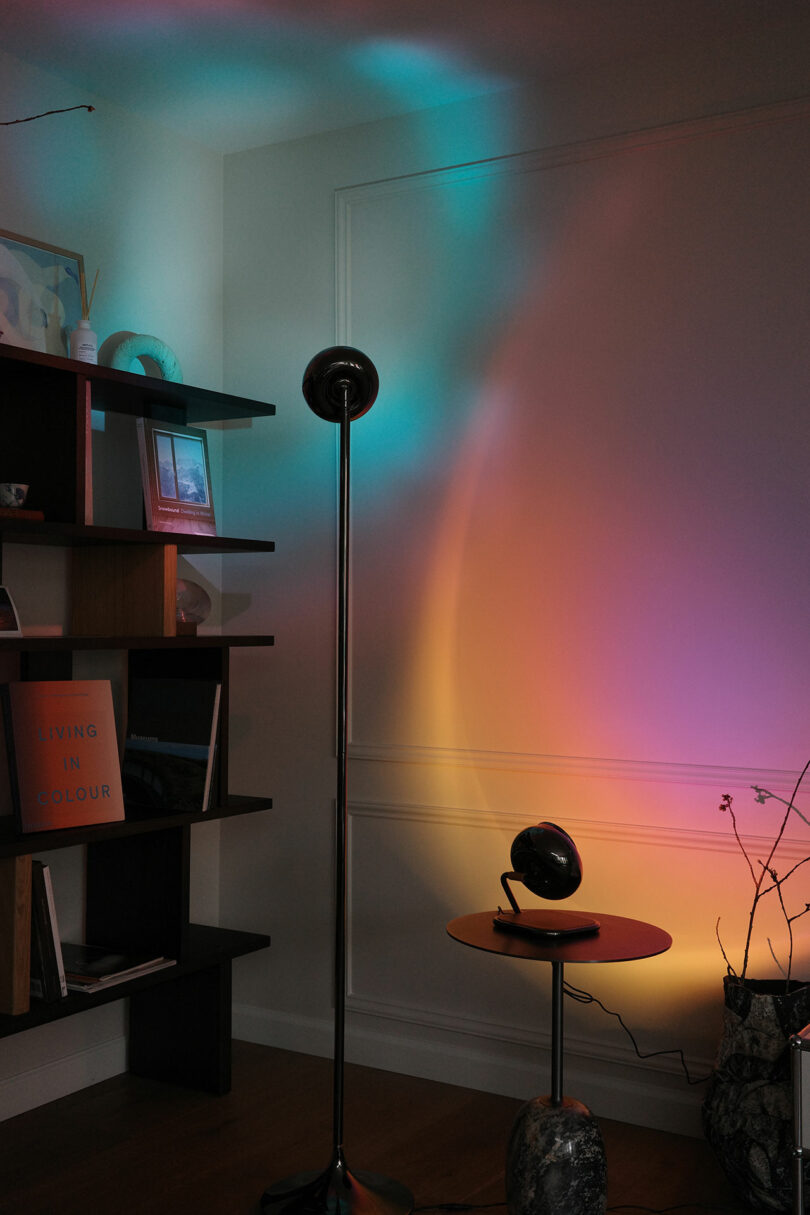 A room corner with colorful lights projected on the wall, a black floor lamp, a small table, shelves with books and decor, and a vase with branches.