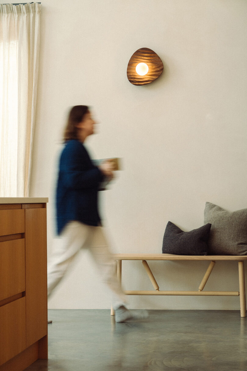 A person walks past a wooden bench with cushions in a minimalist room, holding a mug, with a round wall light above and sheer curtains on the window.