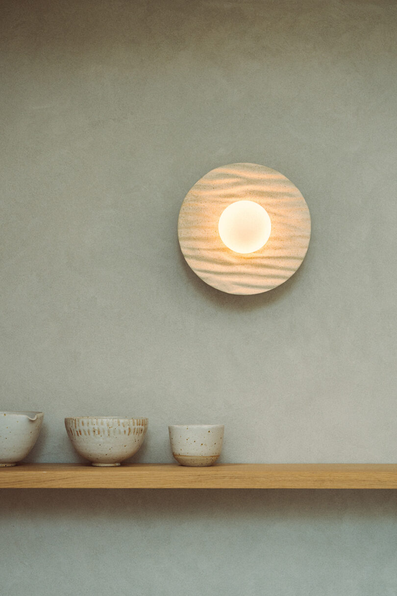 A round wall light is mounted above a wooden shelf holding three ceramic bowls against a textured beige wall.