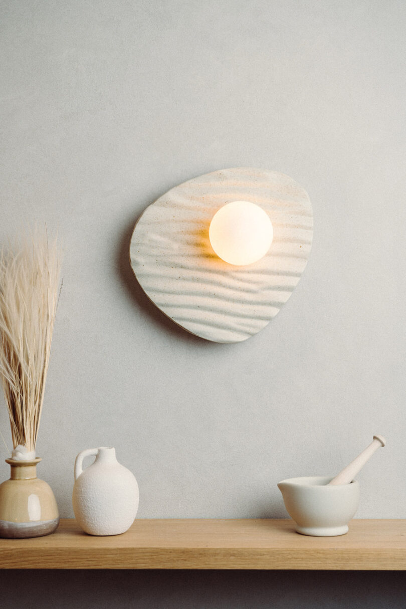 A round wall light with a textured, oval backplate is mounted above a wooden shelf holding dried grasses in a vase, a white jug, and a mortar and pestle.
