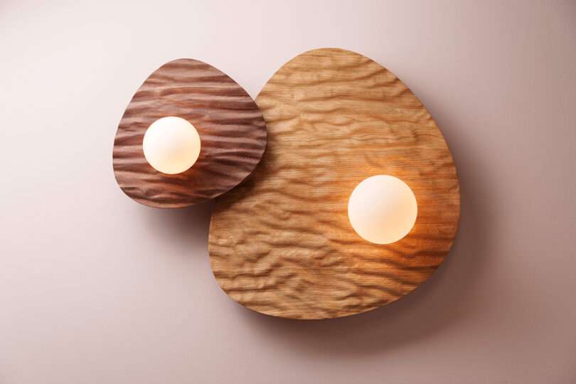 Two round wall lights are mounted on overlapping, irregularly shaped, textured wooden panels against a light-colored wall.