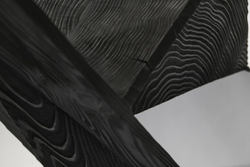 Close-up of blackened wooden beams intersecting at an angle, highlighting distinct wood grain patterns, with a plain light grey background