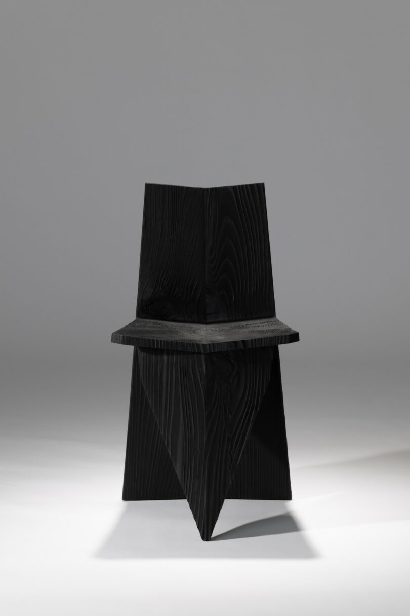 A black, angular wooden chair with geometric shapes and sharp lines is centered against a plain gray background